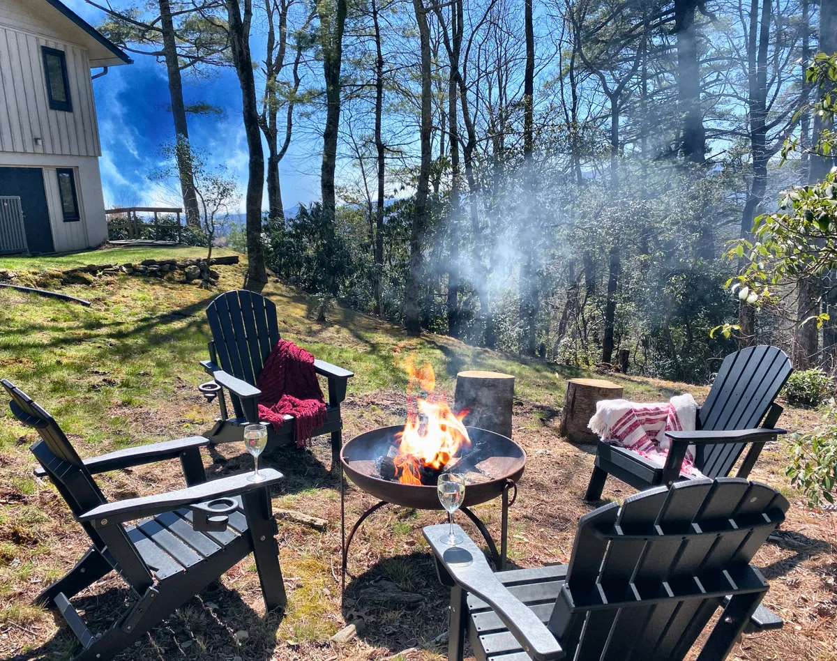 A cozy fire pit area with Adirondack chairs arranged in a circle, wine glasses set on the armrests, and tall trees enclosing the scene. The warm glow from the fire invites guests to gather, while the open sky above hints at opportunities for stargazing and storytelling.