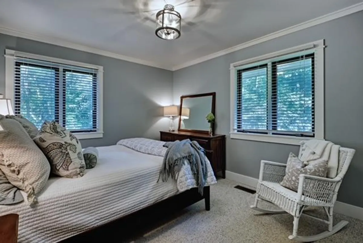 A cozy bedroom highlighted by a comfortable bed, a classic rocking chair beside a lamp, and a large mirror. Natural light streams in, creating a serene atmosphere that's perfect for reading, napping, or quiet retreat.