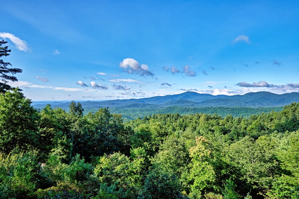 A sweeping mountain view stretches beyond lush green forests under a vast blue sky, with dramatic cloud formations accentuating the natural beauty. The scene is tranquil and inviting, suggesting a peaceful setting perfect for relaxation, exploration, and connection with nature.