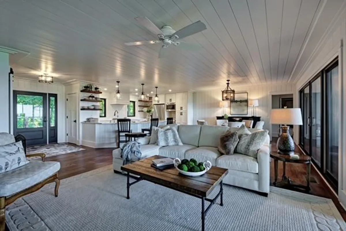 A warm interior scene with a comfortable couch, wooden coffee table topped with greenery, and a dining area visible in the background. Soft natural light streams in, creating an inviting space for gatherings or quiet moments.