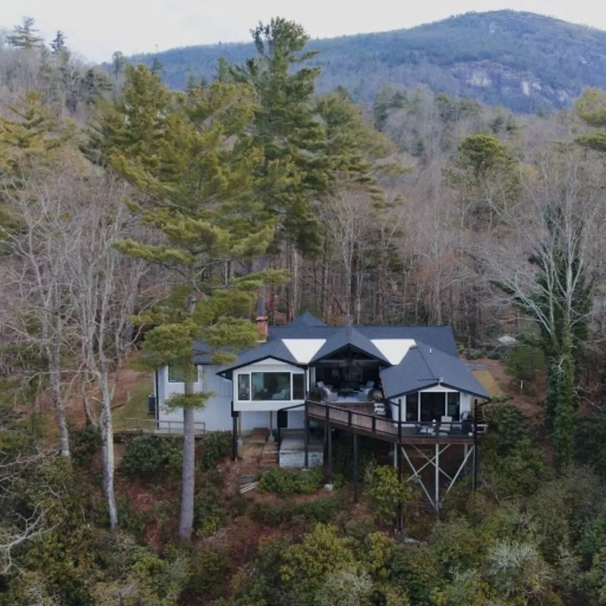 A wide deck extending from the house, surrounded by lush trees and offering a sweeping view of the mountains. The deck is furnished for relaxation and socializing, and the peaceful, natural setting emphasizes the property's scenic location.