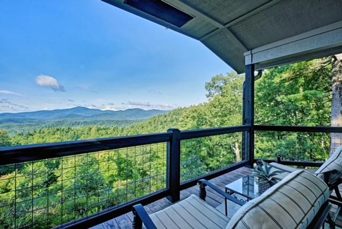 An outdoor seating area with a glass coffee table overlooks a breathtaking mountain vista. The serene setting suggests a perfect spot for relaxing after a day of exploring town, reading, or enjoying a warm drink as the sun rises or sets.