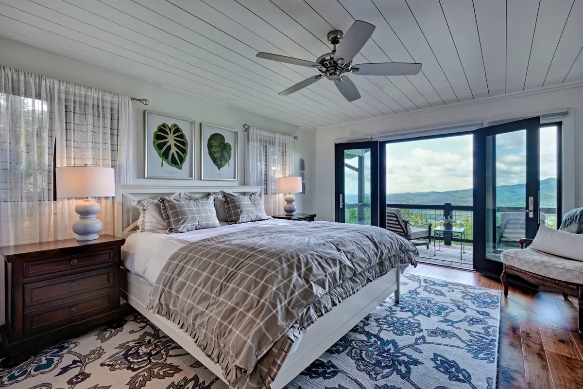 A spacious bedroom featuring a king-sized bed dressed in plush bedding, with large windows providing stunning mountain views. Decorative wall art and bedside table lamps evoke a sense of calm luxury, while natural light fills the space.